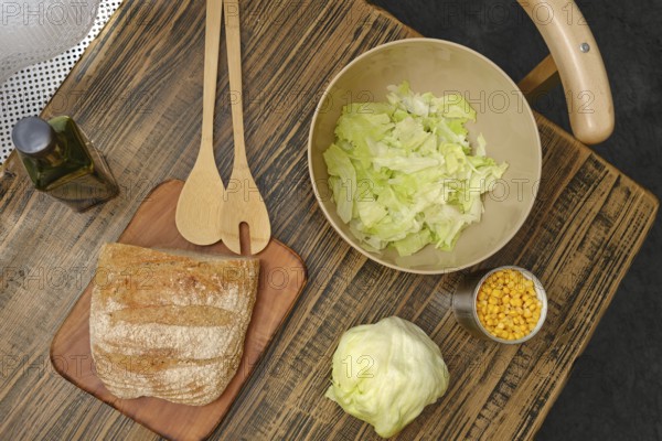 A rustic wooden table displays a loaf of bread, a bowl of chopped lettuce, and a cup of corn. Wooden utensils rest nearby, ready for meal preparation in a cozy kitchen setting