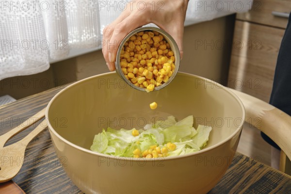 A hand pours canned corn into a bowl filled with fresh green lettuce. The warm kitchen features wood accents, creating a welcoming atmosphere for meal preparation