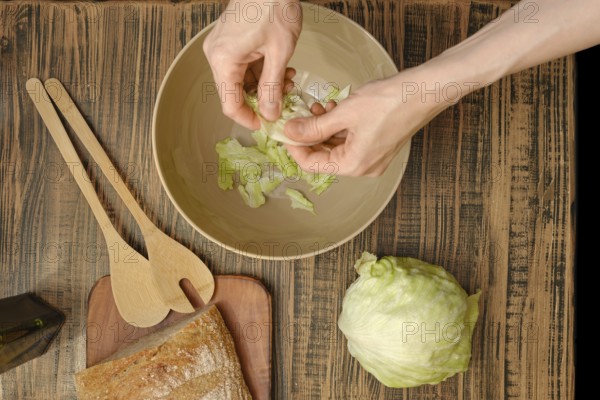 Hands tear crisp lettuce leaves into a bowl, surrounded by wooden utensils and fresh ingredients. The rustic table adds warmth to this vibrant and healthy preparation activity