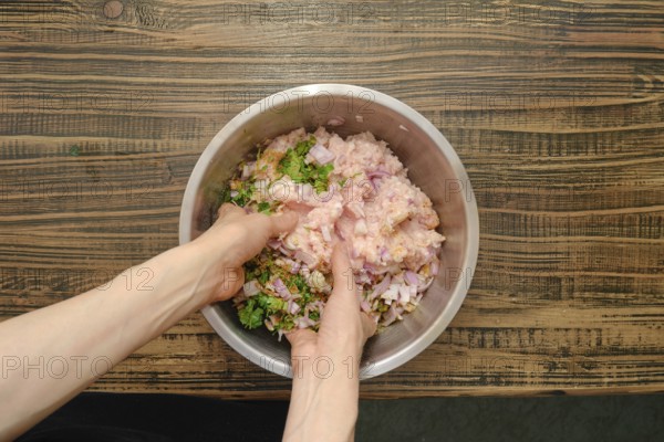 Hands are mixing raw minced meat, chopped onions, herbs, and spices in a stainless steel bowl on a rustic wooden countertop. The preparation indicates a focus on creating a traditional cutlet