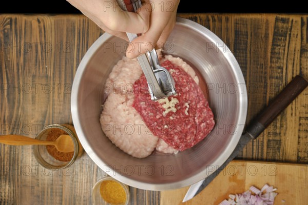 A hands use press to add minced garlic to a mixture of ground meats in a stainless-steel bowl. Various spices are nearby, indicating preparation for a savory dish