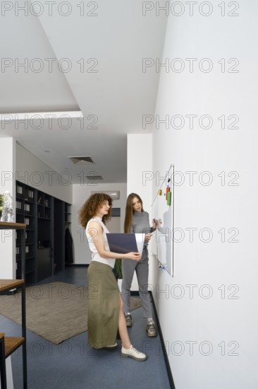 Two young women engage in a brainstorming session in a sleek office, discussing ideas on a whiteboard for their health startup while surrounded by an inspiring and organized workspace
