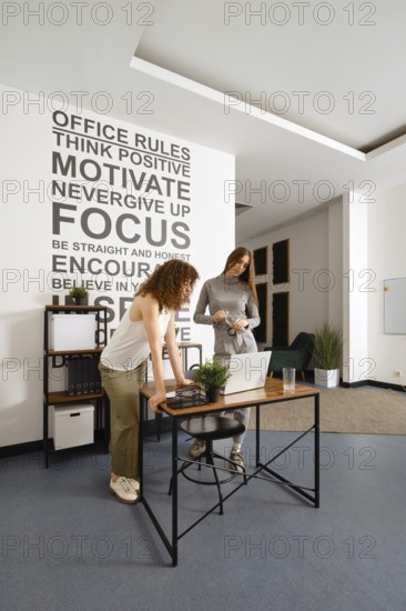 Two women engage in a collaborative work session within a contemporary office environment. The inspiring wall text emphasizes motivation and focus as they discuss women's health initiatives