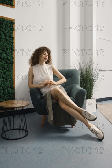 Woman relaxing in a stylish chair in a modern office space with greenery and natural light. She has curly hair and wears a casual outfit suitable for everyday wear