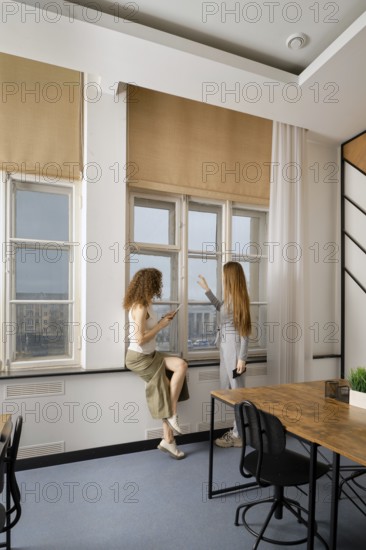 Two women engage in a conversation while standing by large windows in a modern office setting. The atmosphere is professional and vibrant, emphasizing collaboration in women's health initiatives