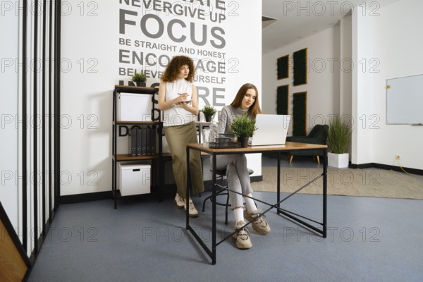 Two women working in bright office. One takes notes while the other types on a laptop, showcasing teamwork and innovative ideas