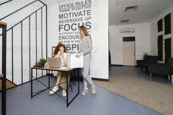 Woman speaks on the phone sitting in coworking and looks at her colleague in surprise, who brought a large box with documents
