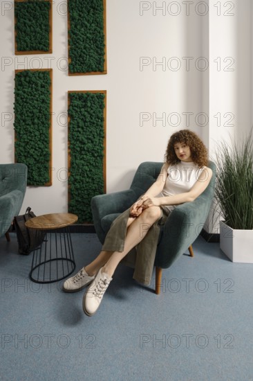 A young woman sits comfortably in a contemporary chair, dressed in a casual outfit. The room features greenery on the walls, stylish furniture, and minimalist decor, creating a serene atmosphere