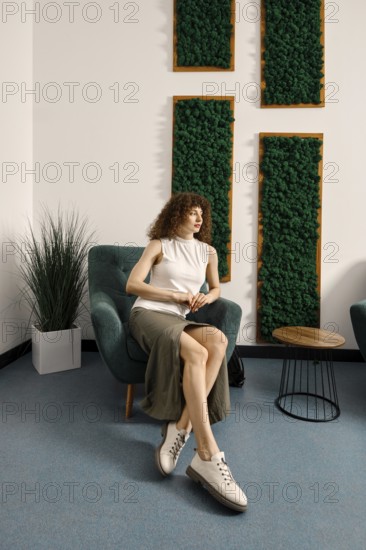 A fashionable woman is seated in a chair within a contemporary room adorned with lush green wall art. She wears a casual outfit
