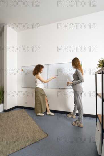 Two women engage in a brainstorming session at a contemporary office. They are standing by a whiteboard for visual organization and idea sharing