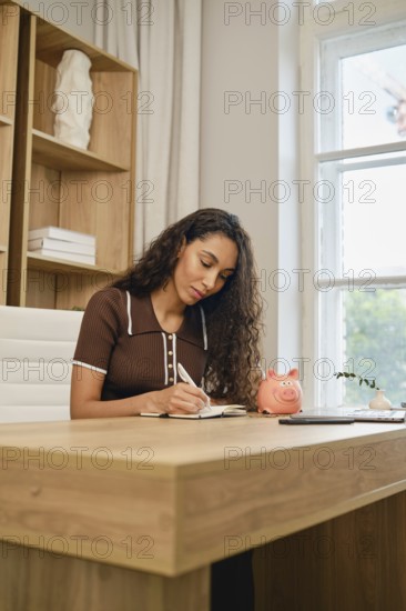 A woman with curly hair is focused on calculating expenses, sitting at a contemporary wooden desk