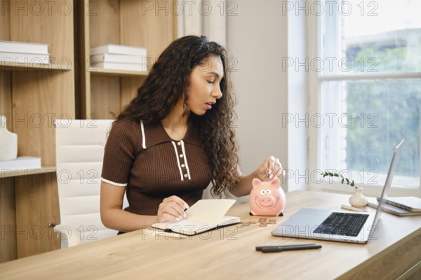 A woman sits at a wooden desk in a stylish home office, writing in a notebook while dropping coins into a pink piggy bank. Natural light filters through the window, creating a warm atmosphere