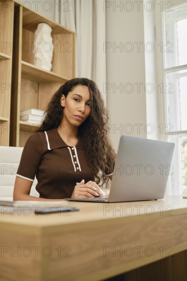 A young woman with curly hair works at the desk using a laptop. She was distracted from the screen and looks thoughtfully at the camera