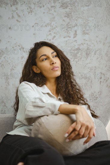 Dreamy woman sits comfortably on a gray sofa, holding a decorative pillow. Her long curly hair cascades down her shoulders as she gazes upward, lost in thought in a stylish interior