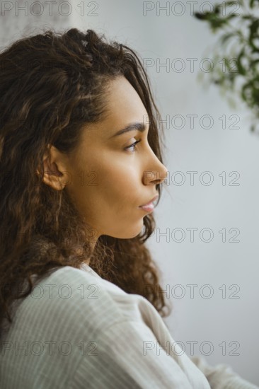 In a cozy indoor space, a woman with long, curly hair sits in profile, lost in thought. Soft natural light highlights her features, creating a serene atmosphere filled with tranquility