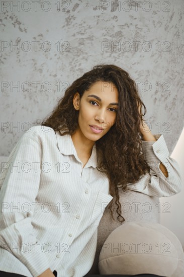 Charming woman with curly hair sits comfortably on a plush sofa in a contemporary indoor space. Sunlight filters through the window, highlighting her relaxed pose and fashionable attire