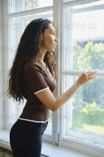 A woman admires the greenery outside while standing by a window. Her curly hair cascades down her shoulders, and she appears contemplative in the warm afternoon light