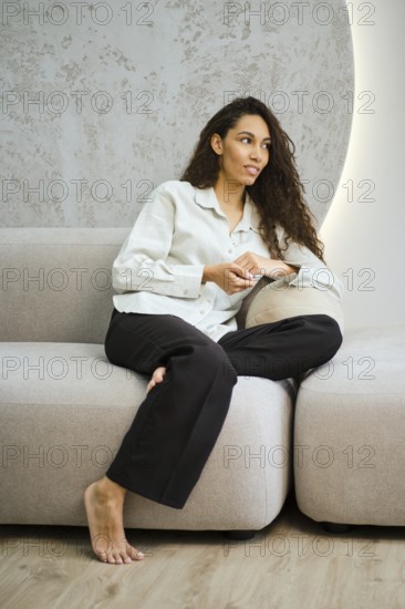 Cute woman with long curly hair sits comfortably on a modern couch in a stylish living room