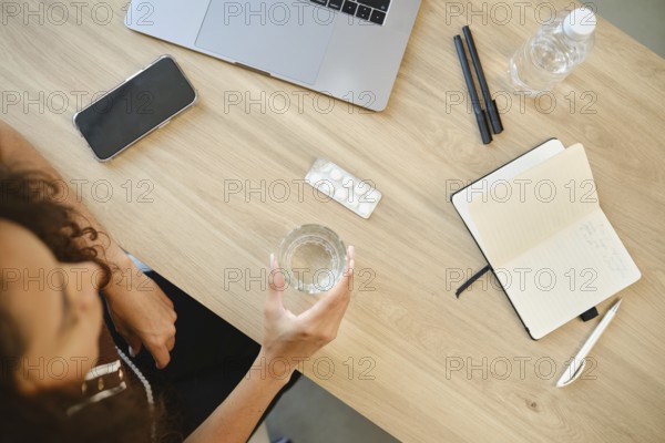 A person sits at a desk with a laptop, holding a glass of water in one hand. A pack of pills and a notebook are visible on a tabletop