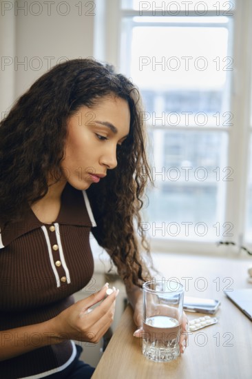 A young woman with curly hair sits at a table concentrating on a pill in her hand and a glass of water beside her. Sunlight filters through the window, creating a serene atmosphere