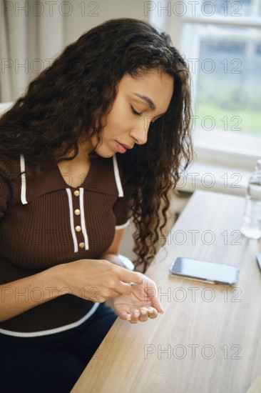 A woman squeezes a pill out of a pack while sitting at a wooden desk in a bright indoor setting
