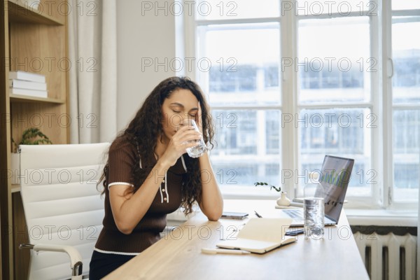 A focused woman sips from a glass of water at her desk during a work session in a well-lit office. She appears deep in thought, reflecting on her tasks while surrounded by paperwork and a laptop