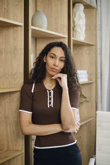 Portrait of a hispanic woman with her hand on her chin, wearing a fitted brown top and black pants. She is surrounded by wooden shelves, contributing to a modern office atmosphere with neutral decor