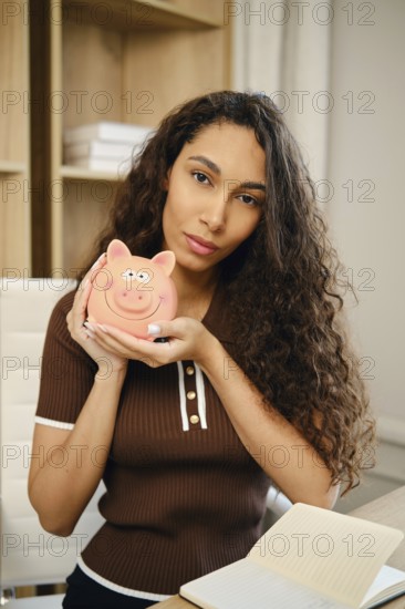 A portrait of a young woman with curly hair holding a cute pink piggy bank. She appears focused and contemplative, suggesting a connection to savings or finance