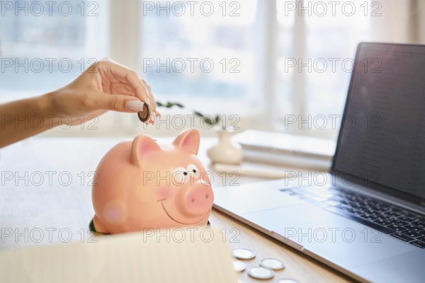 A hand is placing a coin into a pink piggy bank while a laptop sits open on a wooden desk. Sunlight illuminates the tidy workspace, suggesting financial planning