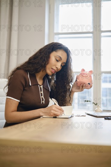 A woman with long curly hair sits at a wooden desk, calculating vacation expenses in a notebook. In one hand, she holds a pink piggy bank