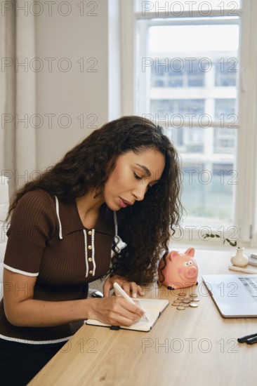 A woman with curly hair writing in her notebook at a wooden desk. She organizes coins beside a pink piggy bank, indicating personal finance management in a bright, cozy room