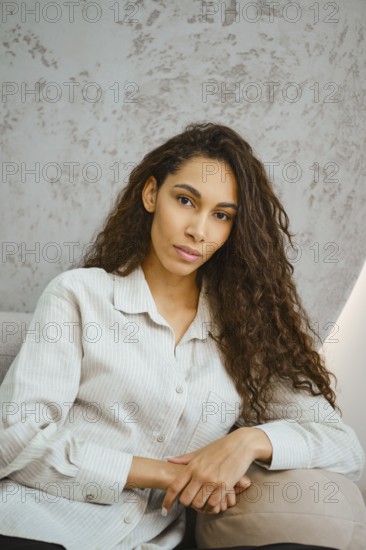 A young woman with long curly hair poses casually in a softly lit modern interior. She rests her arm on armrest of a sofa, exuding a sense of confidence and comfort, dressed in a stylish button-up shirt