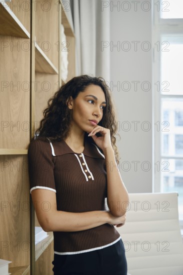A woman with curly hair stands pensively in a well-decorated office. She rests against wooden shelves, deep in thought, as natural light flows through large windows, illuminating her surroundings