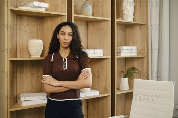A confident businesswoman stands with arms crossed in a contemporary office space. The background features wooden shelves filled with decorative items, creating a stylish ambiance for work