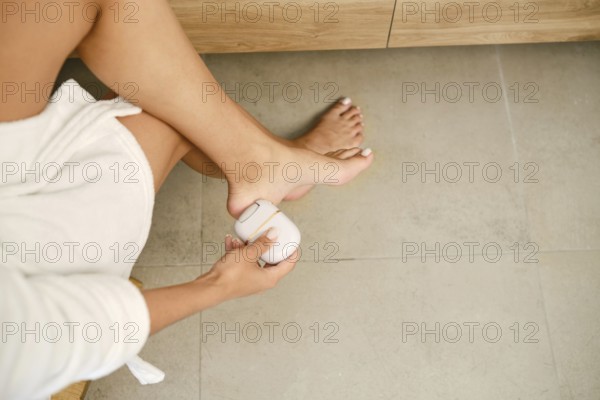 A person sitting on a wooden chair in a bathroom and removing dead skin cells from the outer layer of her feet skin with electric exfoliation machine. Concept of self-care and hygiene