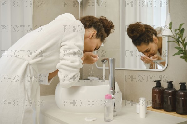 A woman leans over a modern sink, splashing her face with water as part of her skincare routine. She is dressed in a white robe and surrounded by clean, minimalist decor that evokes relaxation