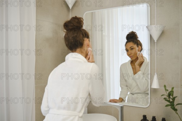 A woman in a white robe is in a sleek bathroom, gently washes makeup off her face. She focuses on her reflection in the mirror, surrounded by elegant decor and soft lighting