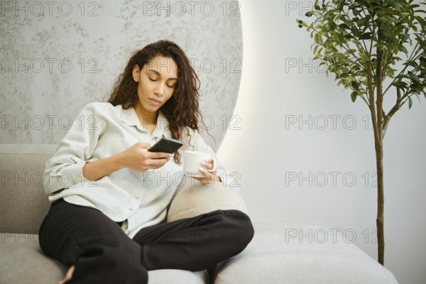 A young woman sits comfortably on a couch in a modern living room, focused on her smartphone while holding a cup. Natural light enhances the relaxed atmosphere of the setting