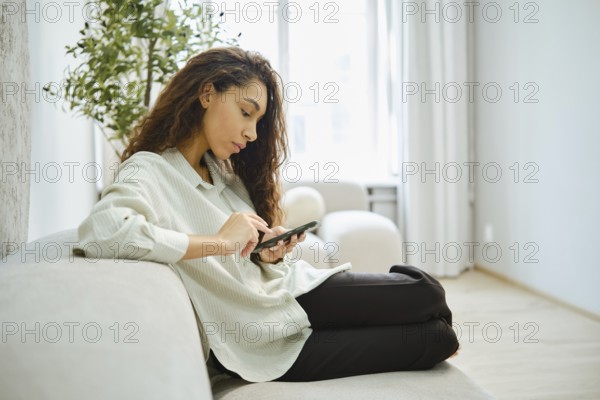 A woman with curly hair sits comfortably on a light-colored sofa, focused on her smartphone in a sunlit living room. A potted plant adds a touch of greenery to the calming space