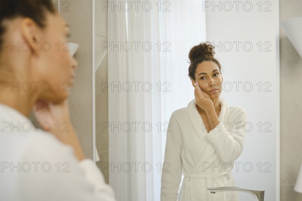A woman in a white bathrobe stands in front of a mirror, applying skincare to her face. The bright, airy bathroom features light-colored decor and natural light filtering through the curtains
