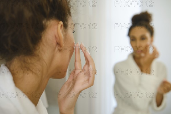 A woman in a white robe is applying cream to her face while looking at herself in the bathroom mirror. Soft lighting creates a serene atmosphere as she follows her morning skincare regimen