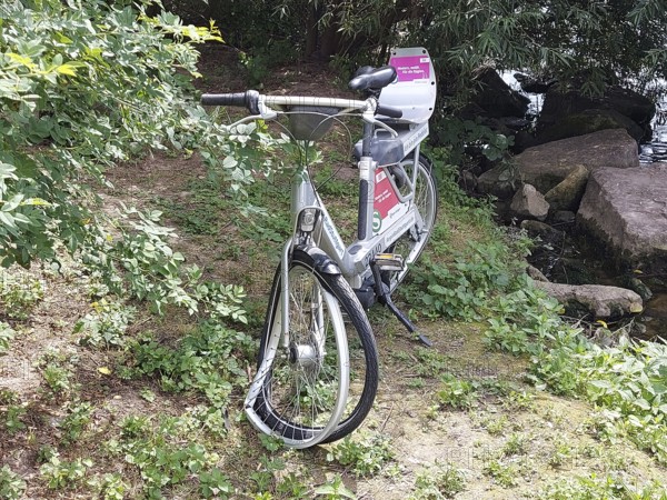 A vandalised bicycle, e-bike from a bike rental company stands in the countryside next to a river, surrounded by trees and stones in a quiet atmosphere, bike sharing, Neckar, Stuttgart, Baden-Württemberg, Germany