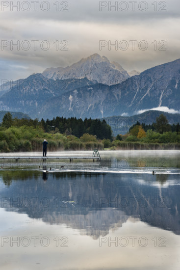 Man (60-70) with dog on a wooden footbridge at sunrise, Hopfensee, near Füssen, Ostallgäu, behind the Tannheimer mountains, Allgäu, Bavaria, Germany