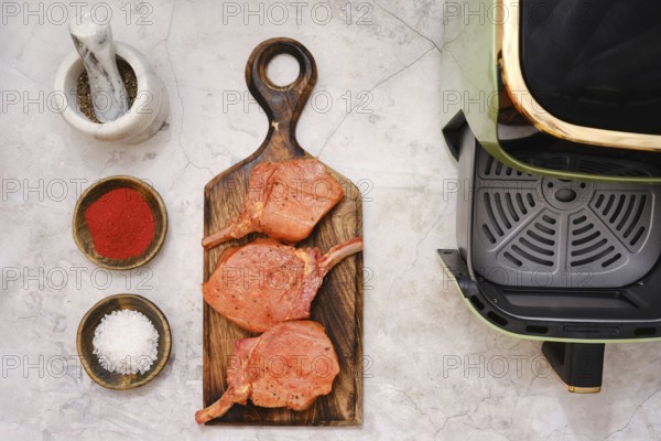 Three pork rib chops are placed on a wooden cutting board, surrounded by spices including paprika and salt in small bowls, and a cooking appliance is visible in the corner, indicating meal prep