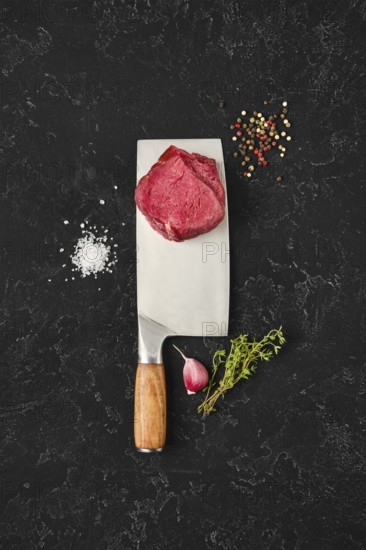 Fresh meat sits atop a large knife on a dark countertop, surrounded by salt, pepper, herbs, and garlic. The setting suggests preparation for a delicious meal, showcasing culinary artistry