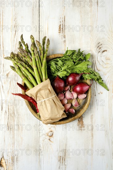 A wooden plate holds an assortment of fresh vegetables and herbs, including asparagus, red shallots, and leafy greens. This vibrant collection is perfect for enhancing culinary dishes or salads