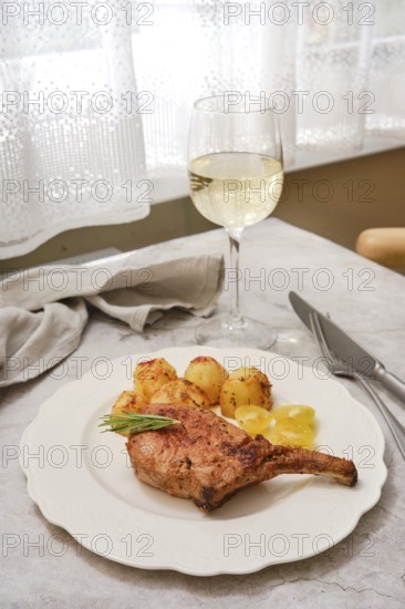 A perfectly roasted pork rib roast is plated alongside crispy golden potatoes and garnished with fresh herbs. A glass of white wine sits nearby, creating an inviting dining atmosphere at home