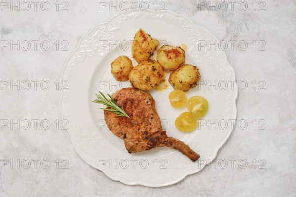 A roasted pork rib roast on a decorative white plate served with golden roasted potatoes and sweet yellow cherry tomatoes