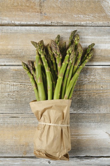 A bunch of fresh green asparagus stalks is wrapped in craft paper and tied with twine, placed on a rustic wooden table. The vibrant green color stands out against the wood