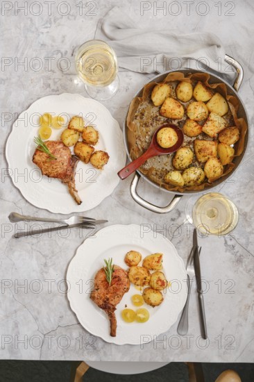 Roasted pork rib roast served alongside beautifully roasted crispy potatoes. The setting features elegant plates and two glasses of white wine, creating a warm dining atmosphere